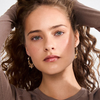 Gold hoop earrings with sculptural charms worn by model with curly brown hair and brown top, front view — Reyca / Boucles d'oreilles créoles dorées avec charms sculptés portées par un mannequin aux cheveux bouclés bruns, vue de face — Reyca