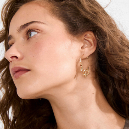 Gold hoop earrings with sculptural charms worn by model with curly brown hair, profile view — Reyca / Boucles d'oreilles créoles dorées avec charms sculptés portées par un mannequin aux cheveux bouclés bruns, vue de profil — Reyca
