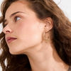 Gold hoop earrings with sculptural charms worn by model with curly brown hair, profile view — Reyca / Boucles d'oreilles créoles dorées avec charms sculptés portées par un mannequin aux cheveux bouclés bruns, vue de profil — Reyca
