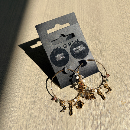 Gold hoop earrings with sculptural charms and glass beads on Pilgrim packaging in natural sunlight — Reyca / Boucles d'oreilles créoles dorées avec charms sculptés et perles de verre sur emballage Pilgrim en lumière naturelle — Reyca