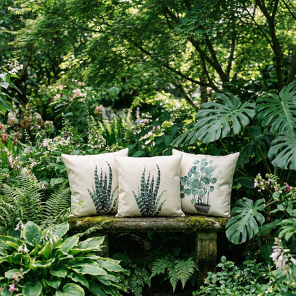 Three botanical print pillow covers on a stone bench in a lush garden, sansevieria and eucalyptus motifs on an ivory background / Trois housses de coussin à imprimé botanique sur un banc de pierre dans un jardin luxuriant, motifs sansevieria et eucalyptus sur fond ivoire