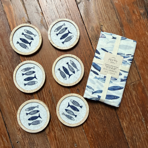Set of six round coasters with fish design on a wooden surface, accompanied by a folded towel with similar design.